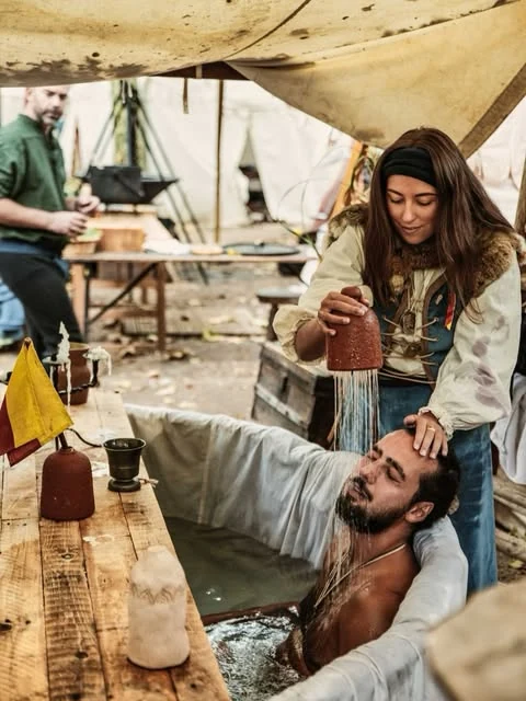 Une compagne en costume tient un pichet près d’un compagnon installé dans un baquet
